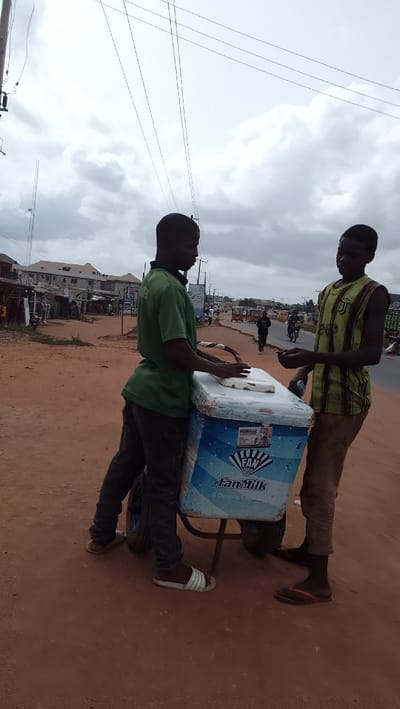 The image depicts a street scene in Ota, Nigeria (latitude: 6.683, longitude: 3.18). Two young men stand on a dirt road, interacting near a mobile cart filled with ice cream. The cart is branded with the "Fan Milk" logo, a recognizable Nigerian dairy product. The man on the left, wearing a green t-shirt, is standing by the cart, while the man on the right, dressed in a striped shirt, is reaching out to him. The scene is set against a backdrop of houses and trees. The sky is partially overcast with fluffy clouds. In the background, there is a blurred image of traffic on the road, indicating a relatively busy street.