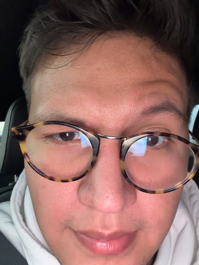 A face, slightly tilted, features a man wearing glasses. The glasses have a tortoiseshell pattern on the frames. The man has brown eyes and dark hair. A small portion of a white garment is visible near his neck. The image appears to be taken from a low angle, focusing primarily on his face and eyewear. The lenses of the glasses reflect light.