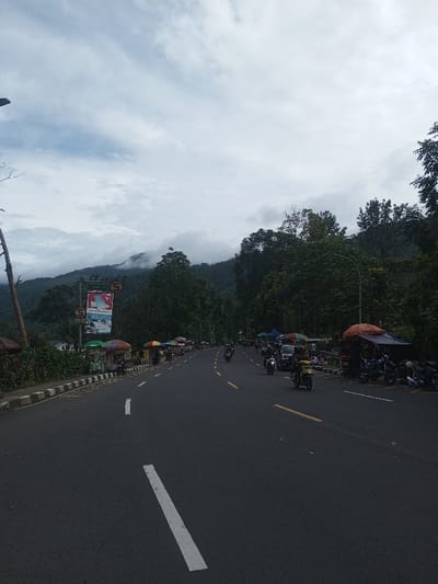 A long, straight asphalt road stretches into the distance, flanked by lush green trees and mountains obscured by low-hanging clouds. Several motorcycles travel along the road, with vendors setting up shop on the sides under colorful umbrellas. Road markings indicate traffic lanes, and a large billboard stands near the roadside. The sky is overcast, casting a diffused light over the scene.