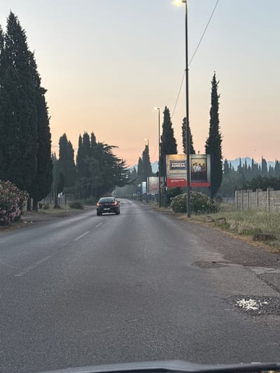 The image depicts a road in Mojanovići, Montenegro, at dawn or dusk, indicated by the soft, pastel colors in the sky. A dark sedan is driving away from the camera down the asphalt road. On the right side of the road, there are several tall cypress trees and a concrete fence. Large billboards are visible along the roadside, one of which displays text in Croatian, reading "JER DOM NIJE SAMO ADRESA" and "STAMBENI KREDITI." A street lamp stands tall near the billboards. The left side of the road is lined with more cypress trees and a large bush with pink flowers. The road surface appears worn, with some potholes visible, particularly on the right shoulder. The overall scene suggests a rural or semi-rural setting on the outskirts of a town.