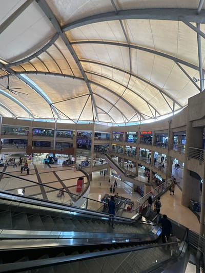 The image depicts the interior of a shopping mall located in Kalyan-Dombivli, India. The mall has multiple floors, visible with various retail outlets and shops. People are seen walking and using escalators. The mall has a large, arched roof structure made of metal supports and a fabric or membrane-like material. Escalators are positioned on the right side of the image. The lighting seems to be a combination of natural light coming through the roof and artificial lighting within the mall.