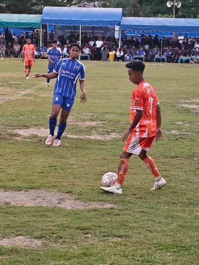 In Desa Meunasah Blang, Indonesia, on a grassy football field, two male football players are actively engaged in a game. The player on the left, wearing a blue and white striped jersey with the number 9 and blue shorts, appears to be moving towards the viewer's right, with his left arm extended. The player on the right, dressed in an orange jersey with the number 7 and matching orange shorts, is dribbling a soccer ball. He is facing the viewer's left, with his right leg positioned over the ball. In the background, spectators are seated under blue and green tents, suggesting a match is in progress. The field itself shows signs of wear, with patches of bare earth amidst the grass.