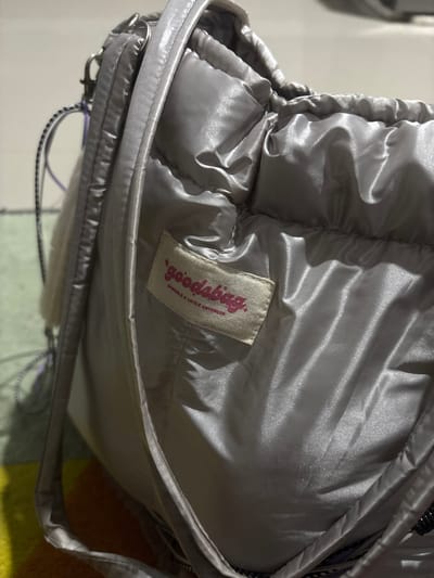 A close-up, indoor, eye-level shot focuses on a silver, puffy bag with a satin-like sheen. The bag's material is gathered and creased, indicating a soft, flexible construction. A small, cream-colored tag with pink lettering is visible on the bag's surface. The lettering on the tag appears to read "goodsbmg" and below it, "SPARKLE & LITTLE CUTE". A strap of the bag is visible along the left side of the frame, attached to a silver clasp. The background is blurred, showing hints of a colorful rug or blanket below and a neutral, out-of-focus wall or surface above. The lighting is artificial and creates strong highlights and shadows on the bag's surface. The image does not provide any information about the city of Tangerang, Indonesia, or any individuals present.