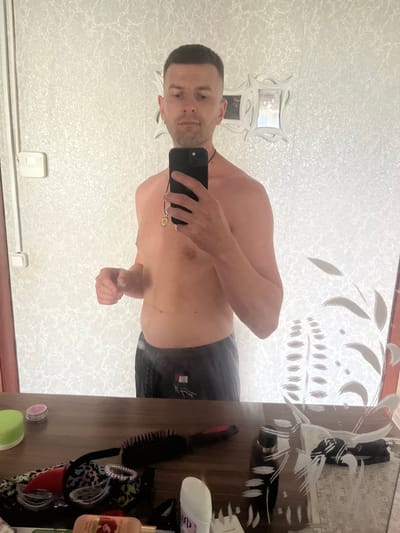 A man in his late 20s or early 30s, with short dark hair and a light complexion, is taking a mirror selfie in a bathroom. He is shirtless, wearing dark grey athletic shorts, and has a small pendant around his neck. His left arm is bent at the elbow, and his right hand is holding a black smartphone. The background consists of a wall with a patterned texture, a light switch on the left, and a decorative mirror with ornate framing. In the foreground, a wooden surface displays various personal items, including a hairbrush, a small container with a pink lid, and other miscellaneous objects. The image appears to have been taken in a residential setting. There is no specific information or indication within the image to suggest the location is in Navahrudak, Belarus.