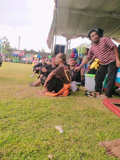 The scene is a grassy field in Karangmalang, Indonesia (latitude: -7.47, longitude: 111.064). There are several men sitting on the grass under a large white canopy tent. They are wearing casual clothing, some with colorful face paint. There are two men sitting on the grass behind the group, also wearing casual clothing. Some are wearing traditional Indonesian clothing. Some men are holding food and drinks, and others are chatting. The environment is bright and sunny, with green grass and a few trees in the background. The men are smiling and laughing. There is a red box on the grass near the group. A white plastic container sits next to the group, with the side missing. There is also a large speaker located behind the tent, but it is not visible in the image.