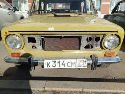 The image shows the front of a yellow, older model car parked on a paved surface. The car has a missing grille, revealing the radiator. The license plate, K 314 CM 29, is visible and reads "RUS" with the Russian flag. The car appears to be in a state of disrepair, with visible rust and peeling paint. In the background, a light-colored SUV is parked to the left, and a darker vehicle is parked to the right. A building with windows is also visible behind the cars. The scene appears to be in an urban environment, consistent with a location in Arkhangelsk, Russia, given the license plate information. The lighting suggests it is daytime. No people are visible in the image.