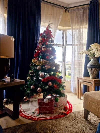 A festive Christmas tree, adorned with colorful ornaments and tinsel, stands near a window. Presents wrapped in festive paper sit beneath the tree on a white and red tree skirt. Blue curtains frame the window and tree. The scene is set on a tiled floor. A table with a vase of white flowers and a woven decoration sits near the window and a chair is visible in the background.