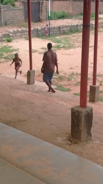 The scene is an outdoor area in Enugwu-Ukwu, Nigeria. There is a man walking barefooted away from the viewer, wearing a plaid shirt and blue shorts. A child is running to his left, wearing a patterned top and shorts. In the background, a child stands near a white flagpole with blue trim. There are three red vertical poles in the foreground, each supported by a square concrete base. A low stone wall borders the background. There is a building and vegetation visible behind the wall. The ground is mostly bare dirt. The foreground appears to be a tiled walkway. A gate is in the background, behind the child at the flagpole.