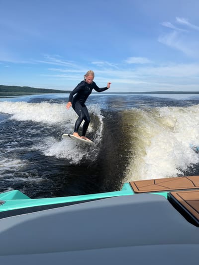 In Gromovo, Russia, a young woman wearing a black wetsuit is surfing on a wave generated by a boat. She is standing on a white surfboard, with her body tilted to the right as she rides the wave. The water is dark and choppy, with white foam from the wave. In the background, there is a forested shoreline under a bright blue sky with scattered white clouds. The front of a boat, with teal and grey accents and brown deck pads, is visible in the foreground.