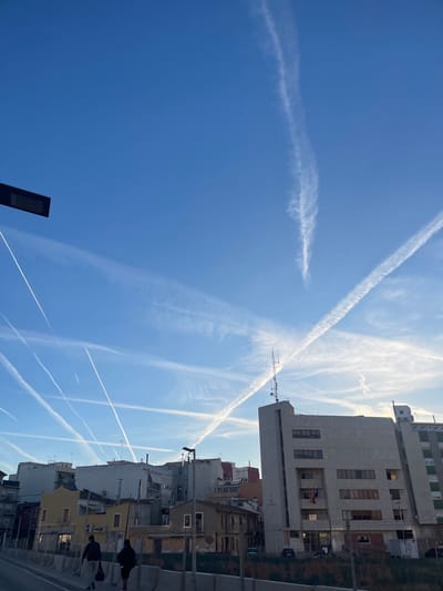 The image shows a view of Paiporta, Spain. The sky is blue with numerous contrails from airplanes. A tall, gray, multi-story building stands in the midground. Adjacent to the building are lower buildings of varying heights, mostly white and yellow. There are people walking on the street. A street light is visible at the left side of the frame.