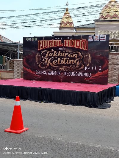 A black stage draped with a red fabric cover is set up in front of a large sign in Anjatan, Indonesia (latitude: -6.41, longitude: 107.961). The sign advertises an event called "Takbiran Keliling Part 2"  hosted by Masjid Nurul Huda. The sign features a dragon with a red, orange, and yellow color scheme.  A single orange traffic cone stands in the foreground. The background features a partially visible mosque building. The area appears to be a street.  The sign is surrounded by electrical wires strung overhead.  A second sign is partially visible to the right, and the remnants of a blue object are visible in the bottom right corner.