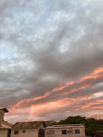 The image captures a sky filled with dramatic clouds, tinged with hues of orange and pink from a sunset or sunrise, set against a backdrop of a pale blue and grey sky. Long, streaky clouds are illuminated from below with a warm, peachy glow, creating a striking visual contrast with the darker, more textured clouds above. A faint rainbow arcs upwards from the lower part of the sky.
In the foreground, several buildings are visible. On the left, a portion of a light-colored building with a sloped, tiled roof is partially seen. To its right, a longer, single-story building with a similar tiled roof is prominent. This building has windows and appears to be a residential or commercial structure. Further to the right, another white building with a flat roof is visible, featuring an air conditioning unit and windows. Behind these buildings, lush green trees form a dense line, and in the far distance, the silhouette of rolling hills can be observed.
The image appears to be taken from an elevated perspective, looking up at the sky and down onto the buildings and landscape. The scene suggests a late afternoon or early morning setting, given the quality of light and the colors in the sky.
While the image itself does not contain explicit geographical markers to pinpoint Florianópolis, Brazil, the description is a general observation of the visual elements present. To confirm the location, additional contextual information would be required.