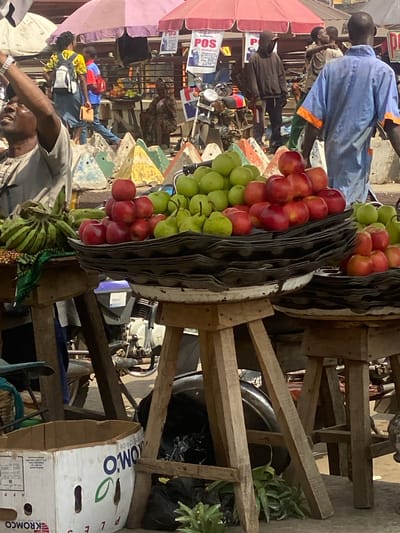 The image portrays a bustling outdoor street market scene in Ibadan, Nigeria. In the immediate foreground, three wooden stalls are laden with produce. The left stall displays a large bunch of green bananas resting on a patterned cloth. The central and right stalls feature multiple stacked black trays, possibly repurposed egg cartons, holding distinct arrangements of red apples, green apples, and green pears. Beneath the left stall, a white cardboard box with partial labels "KROMCO" and "OMOXO" is visible, alongside green plant material on the ground.
Various individuals populate the mid-ground and background. In the left foreground, a man in a light grey patterned shirt gestures upwards with his right arm. Behind the stalls, a woman wearing a yellow patterned shirt, blue skirt, and white backpack walks rightward, carrying a small brown item. A man in a light blue tunic with orange trim on the sleeves walks away from the viewer towards the right. Other observed figures include a man in a dark hooded top, a person in a red and blue shirt, and a seated individual in traditional attire, all engaged in market activities.
Above the scene, multiple large market umbrellas in shades of pink, purple, red, and white provide shade. Banners advertising "POS" (Point of Sale) services, including "Withdraw, Deposit, Transfer, Pay Bills, DATA," are visible under these umbrellas. The ground level features a series of colorful, pyramid-shaped concrete barriers (blue, green, yellow, grey). Several motorcycles are parked or in motion in the mid-ground. The scene is illuminated by bright daylight, indicating an active commercial district.