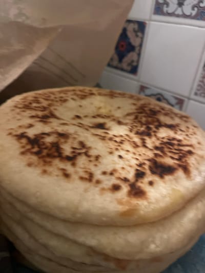 A stack of flatbreads is the focal point, featuring a lightly browned surface with darker speckles. Their soft texture is implied by the visible folds and slight overlapping of each piece. To the left, a nondescript, beige material hints at packaging or a background element. The backdrop is a section of tiled wall, adorned with decorative patterns in blue, white, and red, adding a touch of visual interest. The overall impression is simple and focused on the culinary subject matter.