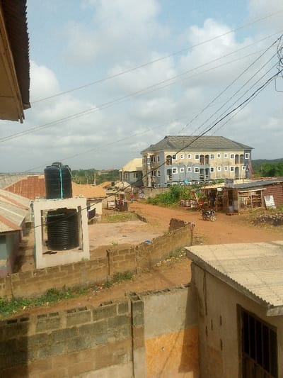 The image depicts an outdoor scene in Alabata, Nigeria, under a partially cloudy sky with visible utility wires overhead.

In the foreground, a low, incomplete wall constructed from unplastered concrete blocks bisects the lower portion of the frame. To the immediate right, the corner of a building with a grey corrugated metal roof and light-colored walls featuring a window with security bars is visible. To the left, a portion of a building with a dark corrugated metal roof overhang is present.

The midground features an unpaved dirt road or path that curves through the scene. On the left, two black plastic water tanks are situated; one is elevated on a concrete block stand with blue piping, and another is at ground level. Behind these tanks, clothes are visible hanging on lines. On the right side of the dirt path, a cluster of rudimentary wooden market stalls or shops with corrugated metal and wood coverings is established. A white motorcycle is parked directly in front of these stalls. Several individuals, including children, are visible walking on the dirt path further into the scene near the shops.

In the background, a prominent multi-story building, approximately four levels tall, stands centrally with a dark grey roof and light blue/grey walls, featuring numerous white-framed windows and balconies with dark railings. A red ladder leans against the right side of this building. Other smaller, single-story and two-story buildings with various roof types are scattered across the distant landscape, interspersed with sparse green vegetation.