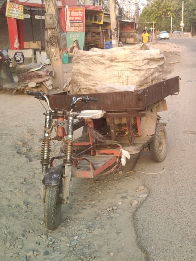 This is a medium shot, taken outdoors, in what appears to be a street scene in Gurgaon, India. The focal point of the image is a three-wheeled electric cart, likely used for transport or deliveries. The cart is loaded with a large, loosely covered bundle that appears to be made of a rough, light-colored fabric. The cart itself is a utilitarian design, with a rusty metal frame, a wooden cargo bed, and visible wiring. It has two wheels at the back and one at the front, which is partially obscured by a mudguard.
The cart is parked on a dusty, uneven surface, with small stones and dirt scattered around. In the background, there are buildings that appear to be shops or businesses, with signs in Hindi, indicating a commercial area. One sign reads "TO-LET" and another has a Bisleri advertisement. An old, possibly damaged scooter is parked to the left of the cart. Further in the background, a man in a yellow t-shirt is standing on the side of a paved road, where a white car is visible. The overall environment suggests a busy, possibly commercial, urban setting. The lighting indicates it is daytime.