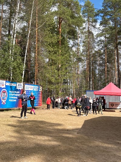 Here's a detailed description of the image:

**General Description:**
The image captures a scene in what appears to be a park or outdoor event setting. There are people gathered, banners displayed, and trees visible in the background.

**Specific Details:**

*   **Location:** Based on the banner content (Russian Federation of Sports Tourism) and the landscape, it's likely in Russia, possibly in Lesnoye, where similar events might occur.
*   **People:** There is a queue of people standing near a building and a red tent. There are two people standinf near the banner.
*   **Objects/Subjects:**
    *   A large blue banner with the words "ФЕДЕРАЦИЯ СПОРТИВНОГО ТУРИЗМА РОССИИ" (Russian Federation of Sports Tourism) is visible.
    *   A scoreboard, probably showing tournament information.
    *   A red tent or canopy set up near a building.
    *   Trees fill the background, typical of a park or forest setting.
*   **Actions/Interactions:** People are standing in line, seemingly waiting for something (registration, information, etc.). The two people near the banner are interacting or posing near the banner.
*   **Environment:** The environment appears to be an outdoor area with a mix of grassy areas and trees. The sky is partly visible, suggesting a clear day.