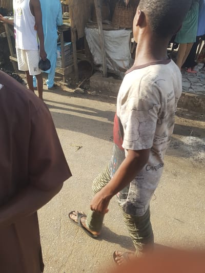 A person walks on a dirt street in Dabawa, Nigeria. The person wears sandals, striped pants, and a patterned t-shirt. Other people are nearby. Buildings of wood and metal are in the background. A brown cloth covers the lower part of the foreground.