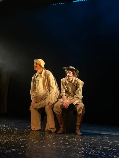 Two male individuals in theatrical costumes are depicted on an indoor theatrical stage in Novoshakhtinsk, Russia. The stage floor is dark, uniformly covered with scattered white particulate matter. Elevated blue stage lights are visible along the upper edge of the frame.
The subject on the left stands, positioned slightly to the right, with his body turned partially left and his head turned sharply right, looking off-frame. He wears a light-toned, long-sleeved shirt, wide-legged trousers, and a matching light-colored cap. A light beige satchel with a flap closure is slung across his right hip. He has a visible mustache, and his mouth is slightly open, suggesting an expression of reaction or speech.
The subject on the right is seated on a dark, indistinct object, facing forward with his head slightly turned left, gazing in that direction. He is attired in a light-colored, long-sleeved shirt, a sleeveless overgarment with multiple dark circular embellishments, light brown breeches, and tall, dark brown lace-up boots. He wears a dark, wide-brimmed hat over dark, curly hair, and possesses a prominent dark mustache and a small soul patch. His hands are clasped together in his lap.
Both subjects are illuminated by directed spotlights from above, casting light primarily on their upper bodies and costumes against a largely black, undefined background. Faint vertical lines, possibly representing stage elements, are visible on the far left of the background.