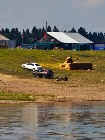 In a rural setting near the city of Pogodaevo, Russia, a body of water occupies the foreground of the image. The middle ground features a grassy slope leading up to a collection of buildings and vehicles. Several trees are visible in the background, with a clear sky overhead.
Dominating the scene is a long, low-slung building with a silver, sloped roof, likely a residential or commercial structure. To its left, a smaller, darker-roofed building with wooden walls is partially obscured. In front of these buildings, a paved or gravel area is visible, along with a fence.
On the grassy slope, two vehicles are parked. A white sedan is positioned higher up, while a dark-colored SUV is parked below it, facing downhill. Behind the SUV, a small, flatbed trailer with wheels is also visible on the slope. Further down the slope, large stacks of firewood are piled on the grass.
The overall impression is one of a remote, possibly rural, settlement with everyday infrastructure. The presence of the water suggests a river or lake, common in the Russian landscape. The weather appears to be fair, with a bright sky.