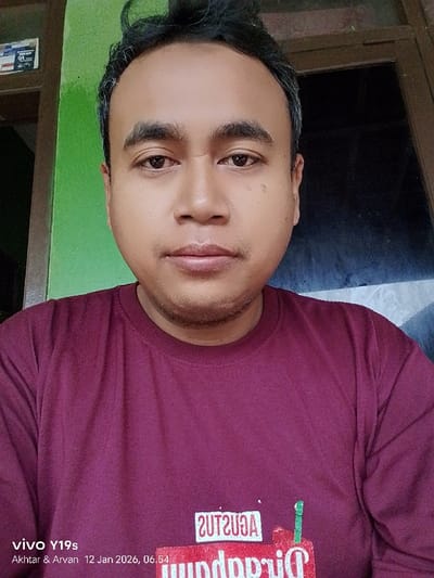 A man poses in front of a green and dark brown background. He wears a maroon t-shirt with a stylized graphic on the lower front, possibly featuring the word "Augustus" backwards. His expression is neutral, and he is positioned centrally in the frame. The photo includes metadata at the bottom left, indicating it was taken with a Vivo Y19s and includes names "Akhtar & Arvan" and a future date.