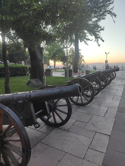The image shows a row of antique cannons lined up along a paved walkway in Osmangazi, Turkey. The cannons are positioned on a slightly elevated area, with a park and trees to the left and a distant landscape visible in the background. The walkway is constructed of large, rectangular stone slabs.

In the foreground, the first cannon is partially visible, with its large wooden wheel and metal barrel prominently featured. The subsequent cannons are lined up behind it, receding into the distance. They are dark metal with wooden wheels.

The park area to the left includes a well-maintained lawn, mature trees, and a hedgerow. In the middle ground of the park, several people are seated at tables and benches. There are also a few individuals walking or standing. In the far background, a hazy sunset is visible, casting a warm glow over the landscape. A lamppost is also present in the background, to the right of the cannons. The overall scene suggests a historical or commemorative site within the city.