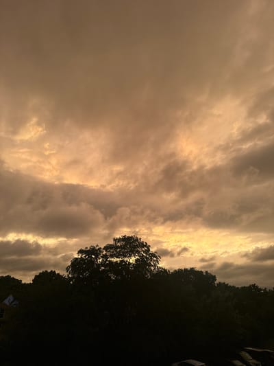 The image captures a dramatic sky over a tree line in Glendale Heights, United States of America. The sky is filled with heavy, textured clouds that are illuminated with a warm, golden light, suggesting either sunrise or sunset. The clouds are dense and layered, with some areas appearing darker and more ominous, hinting at an approaching storm.
Below the sky, a dense silhouette of trees dominates the lower half of the frame. The foliage is dark and appears lush, forming an impenetrable barrier against the light. In the far left of the image, a glimpse of a building with a blue roof is visible through the trees, suggesting the presence of residential or commercial structures within the area.
In the bottom right corner, the dark outlines of parked cars are visible, partially obscured by the trees and the low light conditions. These are the only man-made objects clearly discernible in the foreground. The overall atmosphere of the image is one of natural grandeur and impending change, with the intense sky acting as the focal point.