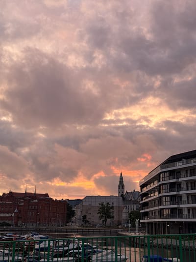 The image captures a sunset over the Oder River in Szczecin, Poland. On the left, a large, red brick building with multiple windows and steep roofs stands prominently. To the right of this structure, a modern, multi-story building with balconies and large glass windows is visible. In the background, a tall, pointed tower with a clock face rises above the cityscape. The sky is filled with dramatic clouds illuminated by the warm hues of the setting sun, ranging from pale orange to pink and grey. In the foreground, a green metal railing separates the viewer from the river, where several boats are moored. The river reflects the colors of the sky.