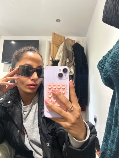 An adult female is captured in a mirror selfie within an indoor room. She wears dark, rectangular sunglasses with silver temple accents, a black puffer jacket over a grey long-sleeved top, and red lipstick. A small stud nose piercing is visible. Her fingernails are painted pink. She holds a light purple smartphone, featuring a pale pink silicone suction-cup grip on its rear case, in her left hand, which also displays a silver ring on the ring finger. Her right hand is raised, with fingers touching the right temple of her sunglasses. In the background, a black flat-screen television is mounted on a white wall to the left. A light brown wooden door, slightly ajar, is visible in the center-right. Behind the door, a coat rack holds several garments, including a light-colored patterned scarf and a dark coat. A dark green textured garment is partially visible on the far right. The room has white walls and a white ceiling with a single recessed light fixture. The location in the city of Winslow, United Kingdom, cannot be determined from the image contents.