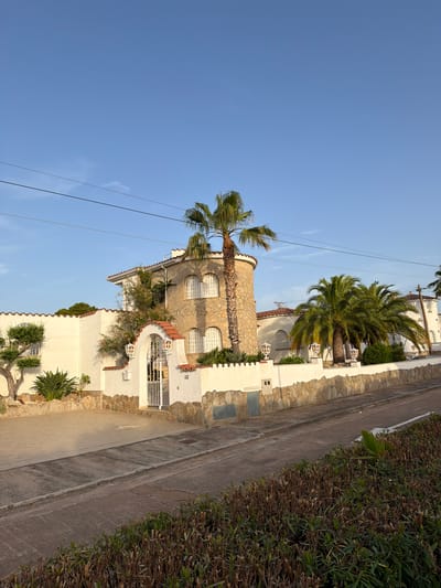 The image depicts a street view in Mont-roig del Camp, Spain, featuring a villa with a white stucco exterior and a stone facade, surrounded by a decorative stone wall. The villa has arched windows and red tile roofing. Lush palm trees and other greenery are visible in the foreground and around the property. A white metal gate provides access to the property, and a paved driveway leads to it. The scene is bathed in the warm light of what appears to be late afternoon or early morning, casting shadows on the road and the facade of the house. The sky is clear and blue, with a few thin clouds. Overhead power lines are also visible, crossing the frame. The overall impression is of a well-maintained residential property in a sunny, possibly coastal, climate.