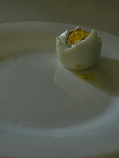 The image shows a close-up of a broken hard-boiled egg with its yolk exposed. The egg is white with a bright yellow yolk that appears slightly crumbly. The egg is sitting in a shallow, white bowl, which is the primary setting. The lighting in the image is dim and somewhat uneven, casting soft shadows and highlighting the texture of the egg. The background is a smooth, white surface, likely the inside of the bowl, with some subtle reflections and a few small yellow specks scattered around, possibly remnants of the yolk. There is no visible text. The scene is simple and focused on the food item. The overall impression is one of a close-up, perhaps a detail shot of food preparation or a meal. The location context of Pyatigorsk, Russia, does not provide any direct visual cues within the image itself.