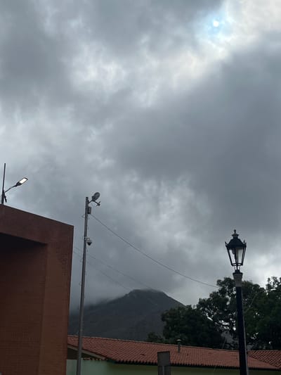 A slightly overcast day is depicted in La Asuncion, Venezuela, with the sun attempting to break through thick clouds in the upper right quadrant. In the foreground, a red brick building occupies the left side of the frame, its flat roofline visible. To the right of the building, a tall utility pole stands, equipped with two surveillance cameras and a street lamp. Further to the right, another lamppost with a decorative, antique-style lantern stands tall. In the mid-ground, a range of trees with dark green foliage is visible. Behind the trees, a mountain shrouded in mist rises, its peak barely visible through the atmospheric haze. The lower portion of the image reveals a tiled roof of a building. The scene is devoid of any visible people or animals.