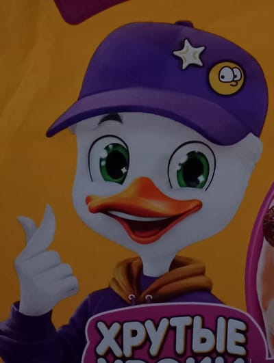The image displays a headshot of a stylized cartoon duck character, rendered in a graphic art style, set against a plain orange background. The duck has a white face, large green eyes with black pupils and white highlights, and an open orange beak showing a red interior, indicative of a smile. It wears a purple baseball cap adorned with two pins: one white star with a yellow outline, and one yellow circular pin featuring a simple cartoon face. The duck is dressed in a purple hooded sweatshirt with an orange hood visible around its neck. Its visible white-gloved left hand performs a "finger heart" gesture, with the thumb and index finger forming a heart shape. Partially visible at the bottom is a pink-bordered purple graphic containing the Russian text "ХРУТЫЕ" in white lettering. This image is observed in Kurganinsk, Russia.