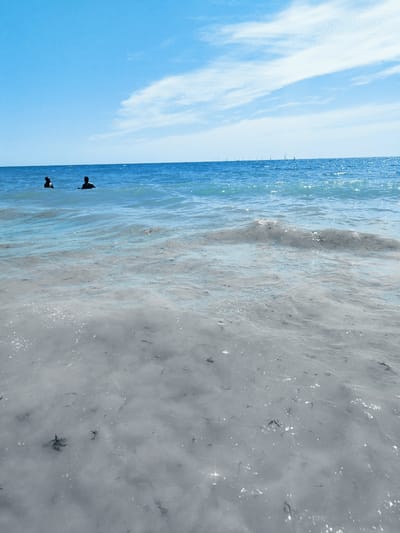 This media file depicts a serene coastal scene at Rota, Spain. The image captures two individuals partially submerged in the ocean, their torsos and heads visible above the water's surface. The water itself is a vibrant blue, transitioning to a lighter, almost translucent hue closer to the shore, where the sandy seabed is visible beneath the gentle waves. Scattered dark specks, possibly seaweed or debris, are present on the seabed.

In the distance, a line of sailboats dots the horizon, suggesting a leisurely day on the water, possibly a regatta or simply recreational sailing. The sky above is a clear, bright blue, with wispy white clouds stretching across it, indicating fair weather. The sunlight glints off the water's surface, creating a shimmering effect. The overall mood of the image is peaceful and idyllic, evoking a sense of relaxation and enjoyment of a summer day at the beach. The time of day appears to be daytime, likely late morning or early afternoon, given the brightness of the sun. There is no visible text in the media file.