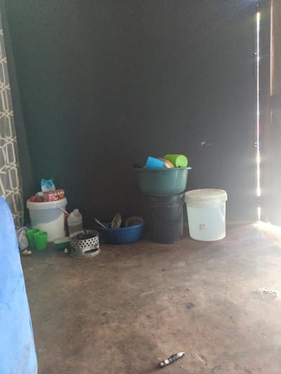 The image, correctly oriented, displays a collection of household or utility items stored in what appears to be a functional indoor or covered outdoor space. On the left, a dark, smooth wall rises, while on the right, a light-colored, stained, and textured concrete or cement floor is visible.

Various plastic containers are prominent. Towards the top, a tall, translucent white plastic bucket stands on the floor. Below it, a dark green bucket is stacked on a black one, holding several smaller, colorful plastic items, including a green cylindrical object and blue pieces. Further down, a blue plastic ring or rim lies on the floor, next to a metallic, perforated cylindrical object, possibly a filter or grate. In the bottom right corner, a white plastic bucket with a red lid is visible, potentially a food product container (faint red text might include "DULAN" or "COOKING OIL"). Another small green cup rests near this bucket. A blue fabric or tarp is partially seen in the bottom right foreground. On the far right of the floor, a small, black and white cylindrical object, possibly a battery or part of a tool, is present. A patterned mat or rug is barely visible along the bottom edge of the dark wall.

Bright light enters the scene from the top, illuminating the floor and objects, suggesting daytime. No people are visible, and no specific activity is occurring; the scene depicts static storage. The utilitarian nature of the setting, with its basic materials and stored items, aligns with a storage or utility area, common in various residential or small commercial properties in Lusaka, Zambia.