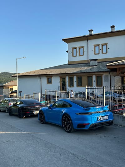 The image captures a street scene in Kerenite, Bulgaria, on a clear day with the sun casting shadows. In the foreground, a vibrant blue Porsche 911 Turbo S is parked facing away from the camera. Its license plate reads "CB 0911 OK". To its left, a black Porsche 911 is parked, with the "PORSCHE" lettering visible on its rear. Further to the left, a silver car is partially visible, followed by a dark-colored van. Behind the cars, a black metal fence encloses a property with a white building featuring brown trim and multiple windows. A person in a white shirt is visible standing near the fence on the left side of the property. The background shows a gently sloping, tree-covered hill under a clear blue sky with a faint contrail. The road surface is asphalt, and a lamppost stands to the left of the parked vehicles.