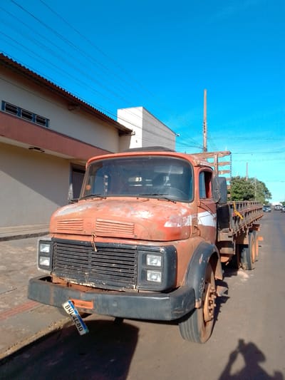 This is a medium-shot, eye-level, outdoor, daytime photograph taken on a street in São Gabriel do Oeste, Brazil. The primary subject is a weathered, orange flatbed truck with a wooden cargo bed, parked facing forward. The truck's paint is peeling and rusted, revealing signs of age and use. A white stripe is visible on the side of the cab. The truck's front bumper is dark gray and appears damaged. A license plate, reading "BWL4E8G", is hanging loosely from the front of the bumper. The truck's windshield is dusty.

Behind the truck, a light-colored building with a reddish-brown trim and several small windows runs along the left side of the frame. Further back, a white building is visible. Power lines stretch across the bright blue sky above. The street appears to be paved, and there is a sidewalk area to the left of the truck. In the background, trees and other vehicles are visible on the street, suggesting this is an urban or semi-urban area. The lighting indicates a sunny day, with shadows cast on the ground, including the shadow of the photographer. The overall impression is a snapshot of everyday life in a Brazilian town.