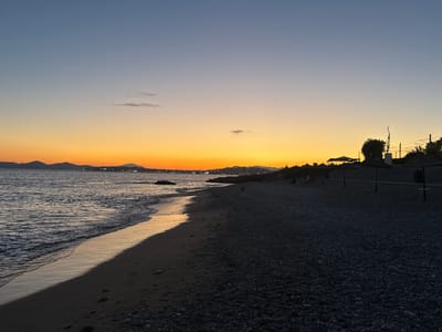 The image depicts a beach at dusk in Paleo Faliro, Greece. The sky transitions from a deep blue at the top to a vibrant orange glow near the horizon, indicating the sunset. The sea, with gentle waves, laps onto a dark, pebbly beach in the foreground. The shoreline is visible, wet and reflective from the receding tide, with footprints suggesting recent activity. In the mid-ground, silhouetted against the sunset, are several thatched umbrellas, likely part of beach amenities, and a distinctive abstract sculpture. Beyond these, more distant hills and lights suggest the urban landscape of Paleo Faliro. The overall atmosphere is serene and tranquil, typical of a beach setting at the end of the day.