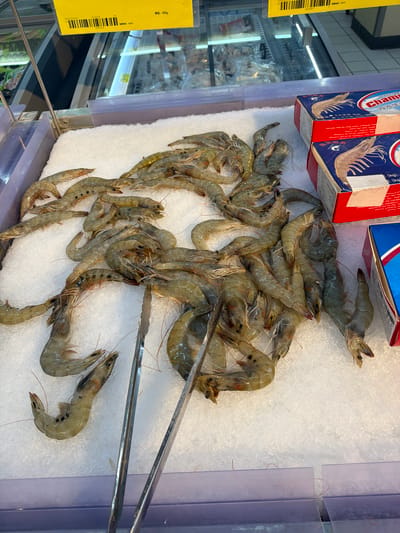 The image shows a refrigerated display of fresh shrimp at a market, presumably in Yuhuatai District, China, based on the Chinese labeling. The shrimp are piled on a bed of crushed ice in a stainless-steel tray with clear plastic dividers. A pair of silver metal tongs is also present. Boxes of Chani-branded shrimp are visible at the upper left. A yellow price tag with a barcode and Chinese text is attached to the left side of the display. The refrigeration unit behind the display contains more prepackaged goods.