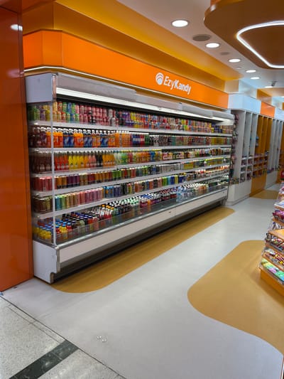 This image captures the interior of an EzyKandy store in Indooroopilly, Australia, showcasing a vibrant and organized display of confectionery and snacks. The scene is presented from a slightly elevated, oblique angle, looking down a long aisle.

The primary subjects are multiple long, glass-fronted display cases, densely packed with a colorful variety of packaged candies, chocolates, and possibly drinks, neatly arranged in vertical columns. These displays extend along the left side of the frame, indicating an extensive selection. Behind and above these main displays, shelves with additional products are visible, though less filled.

The store's branding is prominent, with "EzyKandy" written in white lettering alongside a stylized logo on a bright orange wall panel that runs along the left. The interior design features a striking color palette of orange, light grey, and mustard yellow. The walls are predominantly orange, with recessed circular lights illuminating the space. The floor is a distinctive light grey or white, adorned with large, flowing, organic shapes in mustard yellow, creating a playful and inviting pathway.

No people are present in the image, and no specific activity is depicted, though the setup clearly indicates a retail environment designed for browsing and purchasing. The bright interior lighting suggests the store is open for business, regardless of the time of day or outdoor weather.