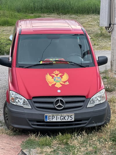 This media file shows a red Mercedes-Benz Sprinter van parked outdoors on a grassy area next to a paved surface. The van is adorned with a prominent coat of arms on its hood, which appears to be the national emblem of Montenegro. The license plate is visible and reads "EPI GK75" with a Polish flag emblem. The scene is well-lit, suggesting it is daytime, and the surrounding vegetation indicates an outdoor setting, possibly a rural or semi-rural location. The orientation of the image is such that the van appears to be on its side, but the details of the vehicle, its emblem, and the license plate are clear. No people are visible in the image. The setting is consistent with Vranjina, Montenegro, given the presence of the Montenegrin coat of arms. The weather appears to be clear and sunny.