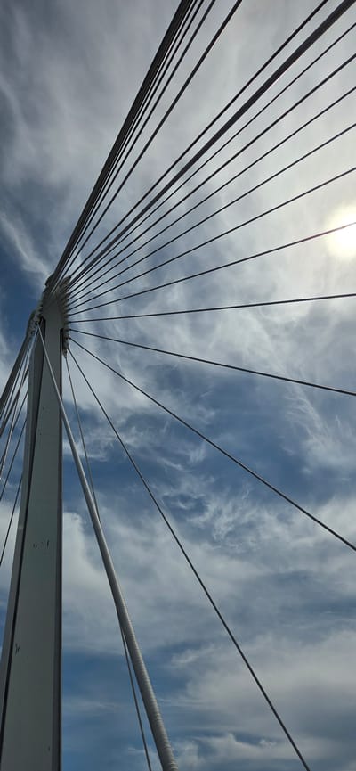 A low-angle shot captures the stark white tower and radiating steel cables of a cable-stayed bridge against a dynamic sky filled with wispy white clouds and patches of blue. The sun is partially obscured by clouds, casting a bright glow in the upper right quadrant. The bridge structure is prominent on the left side of the frame, with numerous thin cables extending upwards and to the right. The scene is devoid of people or any other discernible objects or activities. This perspective focuses on the architectural design of the bridge against a natural backdrop.