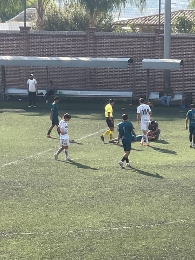 The image depicts an outdoor soccer (football) match or training session paused on an artificial turf field in León, Mexico.
The scene is dominated by a green artificial turf field marked with white lines. In the foreground and mid-ground, several individuals are present. One player, wearing a white jersey with the number "189" on the back, stands upright. Another player in a white jersey with the number "15" on the front walks towards the left. A referee, dressed in a yellow shirt, black shorts, and yellow socks, walks towards the right. One player, wearing a teal/dark green jersey with the number "7" on the back, walks towards the foreground. Another player in a similar teal/dark green jersey with the number "25" is partially visible on the far right.
Central to the mid-ground, a player lies prone on the ground while another individual, wearing a dark top and shorts, kneels beside them, appearing to provide assistance. Two small orange cones are visible on the turf near the referee.
Along the far side of the field, there is a red brick wall. In front of the wall, a long, metal-framed dugout structure with a light-colored, curved roof provides shelter over a bench. Under the left section of the dugout, one person in a white t-shirt stands, and two others are seated on the bench. Under the right section of the dugout, one individual is seated alone, looking at a device.
Above the brick wall, green foliage, including palm trees, is visible. In the distant background, a building with a reddish-brown tiled roof can be seen. The scene is illuminated by bright daylight, casting distinct shadows of the people and structures on the field.
