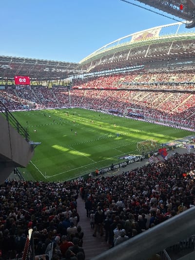 This image depicts a wide view of a professional soccer stadium during a daytime match. The location is identified as the Red Bull Arena (also known as Zentralstadion) in Leipzig, Germany, characterized by its distinctive curved, translucent roof structure visible against a clear blue sky.
Thousands of spectators are present, occupying multiple tiers of seating; a significant portion of the crowd in the visible stands wears red attire. On the bright green, well-maintained soccer pitch, approximately fifteen to twenty players in dark and light athletic uniforms are distributed across the field, suggesting active play or pre-game/post-game presence. A large digital scoreboard in the upper left section of the stadium displays "0:0" in red text, alongside what appears to be "RBL" (likely referencing RB Leipzig). A white goal structure is visible on the right side of the field. The foreground shows a dark metal railing and concrete stadium structure, indicating the viewer's position within the upper stands. Sunlight illuminates the field and portions of the stands, casting visible shadows.