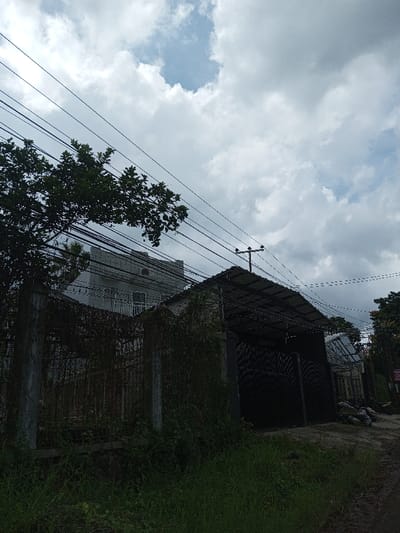 An exterior daytime scene in Sukanagalih, Indonesia, under an overcast sky with scattered patches of blue visible through dense grey clouds. The foreground consists of overgrown grass and weeds, transitioning to an unpaved dirt path on the far right.
In the midground, a light-colored, multi-story residential building featuring a second-floor balcony and an exterior air conditioning unit is partially visible on the left, set behind a heavily overgrown metal fence supported by concrete posts. Adjacent to this on the right is a dark, single-story structure, likely industrial or a large garage, characterized by a dark corrugated metal roof and large, closed dark gates. A utility pole equipped with multiple cross arms and electrical insulators stands atop the roofline of this dark building. Numerous dark utility lines crisscross the upper half of the frame, connecting to the pole. A dense green tree occupies the upper left quadrant, partially obscuring the sky and utility lines. Piles of refuse or debris are situated on the unpaved path next to the dark building on the right. No individuals are present in the scene.