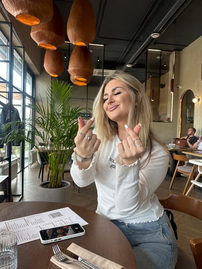 A blonde woman is seated at a dark wooden table within a modern restaurant or cafe in Усады, Russia. She wears a white long-sleeved top with ruffled edges and light blue jeans. Her eyes are closed, and she exhibits a slight smile while forming a finger heart gesture with both hands. Her right hand displays a heart-shaped ring and a thin band, while her left hand wears multiple rings and a square-faced watch. Manicured nails are visible, white on the left hand and light peach on the right. On the table, a menu with a QR code and Cyrillic text is present, alongside a white smartphone displaying an image of the restaurant's hanging lamps, a clear glass of water, and silverware resting on a beige napkin. The restaurant's interior features several reddish-brown, textured, bell-shaped hanging lamps, a black ceiling with exposed ventilation ducts, large windows on the left, and a large potted green plant. Two male patrons are visible, blurred, at a table in the background to the right, and an arched mirror with wall sconces adorns the right wall.