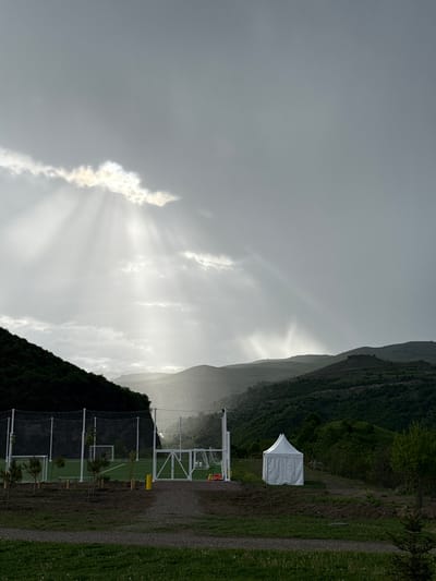 The image shows a landscape with a mountain range in the background. The sky is overcast with diffused sunlight breaking through the clouds. In the mid-ground, there's a green field, possibly a sporting area, featuring white fencing, what appears to be trees, and a small white tent. The location may be in the city of Chkalov, Armenia, but the information cannot be verified.