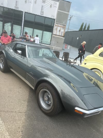 A grey Chevrolet Corvette Stingray is parked outdoors on an asphalt surface. The car is the primary subject, with its side profile prominently displayed. The iconic "Stingray" badging is visible on the car's fender. In the background, a yellow classic car, possibly a Volkswagen Beetle, is also visible. Several people are present, some standing and others seated, suggesting an outdoor gathering or car show. One person in the background is wearing a red and white patterned shirt. The setting appears to be a street or parking area adjacent to a modern building with large windows, which is partially covered with construction sheeting bearing text such as "HAYDUK LUMBER AND HARDWARE" and "TITAN." The sky is overcast, indicating a cloudy day. The time of day is difficult to ascertain precisely, but the lighting suggests it is daytime. The presence of multiple classic cars and onlookers implies an event related to car enthusiasts.