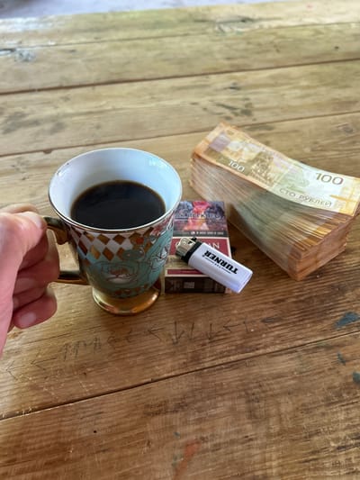 A hand is holding a mug of coffee. The mug is decorated with a pattern of green roses and gold diamonds, and has a gold rim and handle. To the right of the mug, there is a pack of cigarettes, a white lighter with "TURNER" printed on it, and a large stack of Belarusian 100 ruble banknotes. The scene is set on a wooden table with visible grain and some scratches. Some directional arrows are scratched into the table surface. The environment suggests an outdoor or rustic setting.