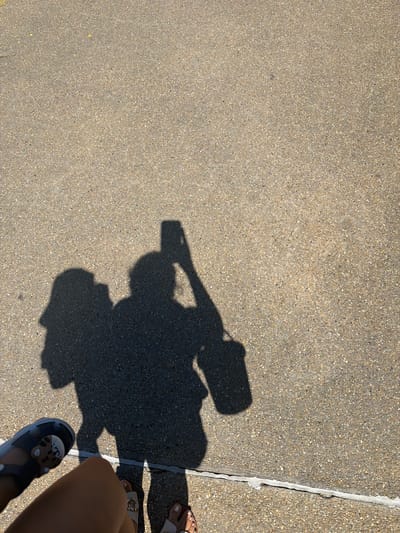 The image is a top-down view of a shadow of two people on a textured, light brown, concrete surface. The person on the left is holding a phone up to take a photo, casting a shadow of their arm and the phone. The person on the right is slightly behind and to the right of the first person, and their shadow is less defined, with a bucket-like object casting a shadow near their hand. In the foreground, the legs and feet of the person taking the photo are visible. They are wearing a grey sandal with white and black embellishments on the left foot, and their right foot is bare, showing painted toenails. The concrete surface has a fine aggregate texture and is marked by a thin, light-colored line running horizontally across the bottom of the frame, with some repair material visible along it. The scene appears to be outdoors on a sunny day, casting strong shadows. The location is Pervolia, Cyprus.